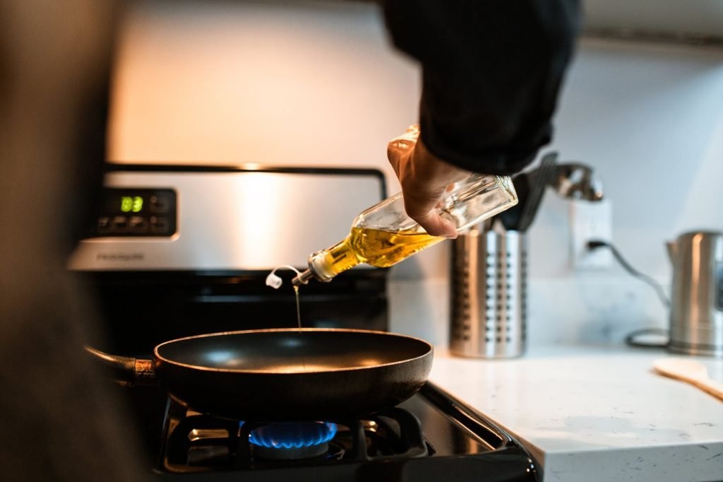 Back view crop unrecognizable person pouring olive or sunflower oil into frying pan placed on stove in domestic kitchen